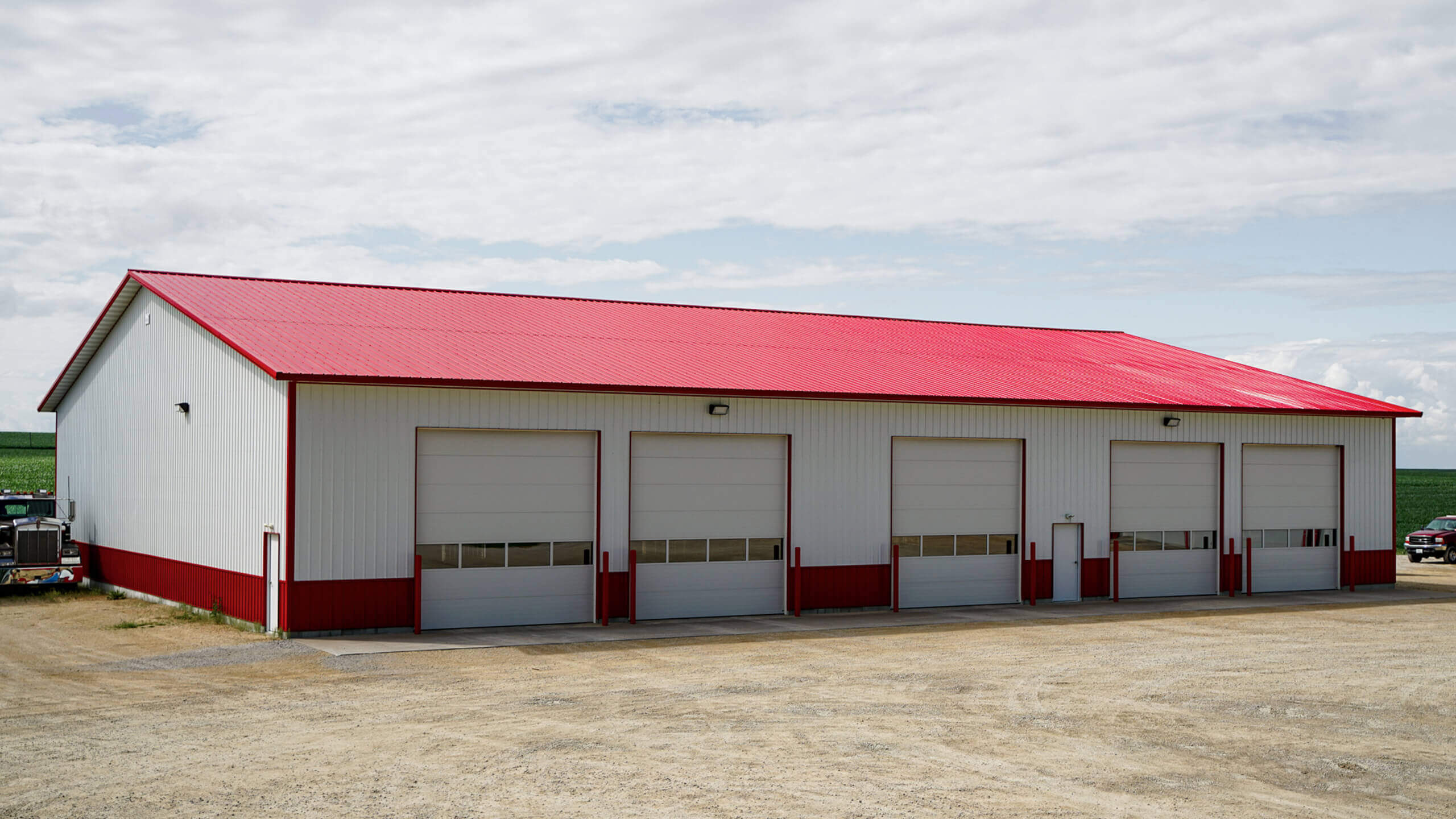 A large white building with a red roof and four closed garage doors sits on a gravel lot under a partly cloudy sky.