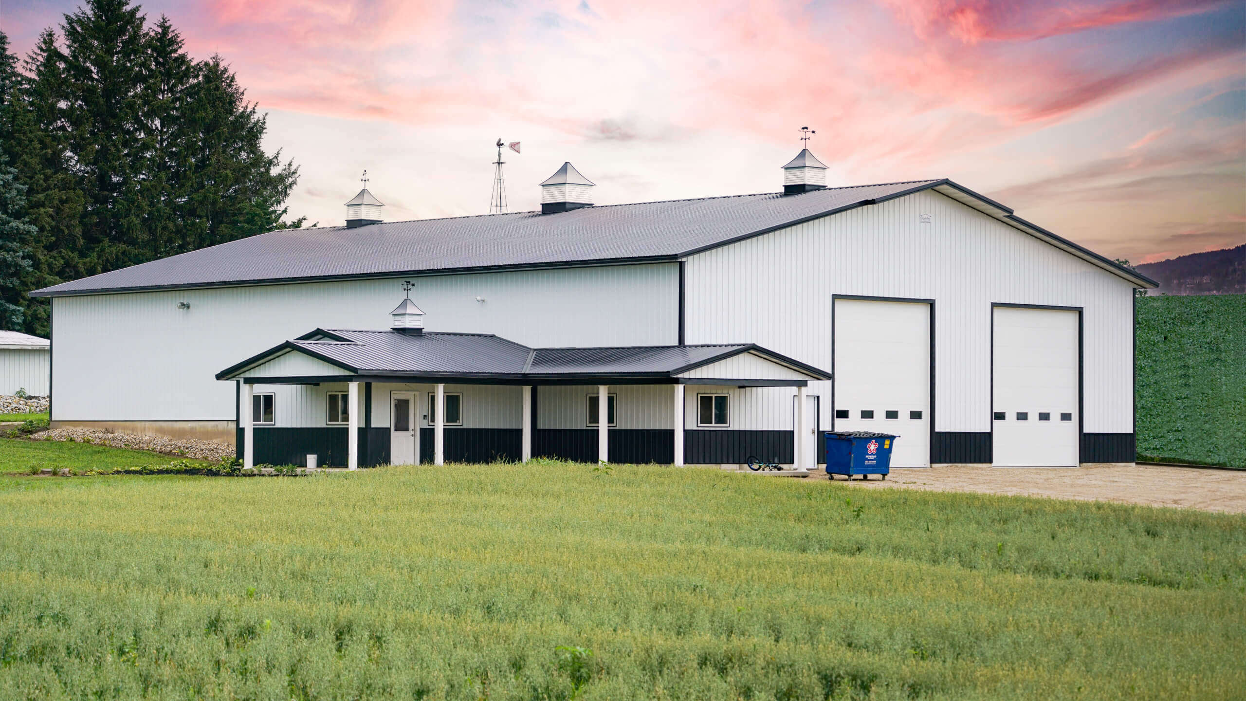 Large white and black barn with three cupolas, two large garage doors, and a dumpster outside, set behind a green field under a colorful sky.