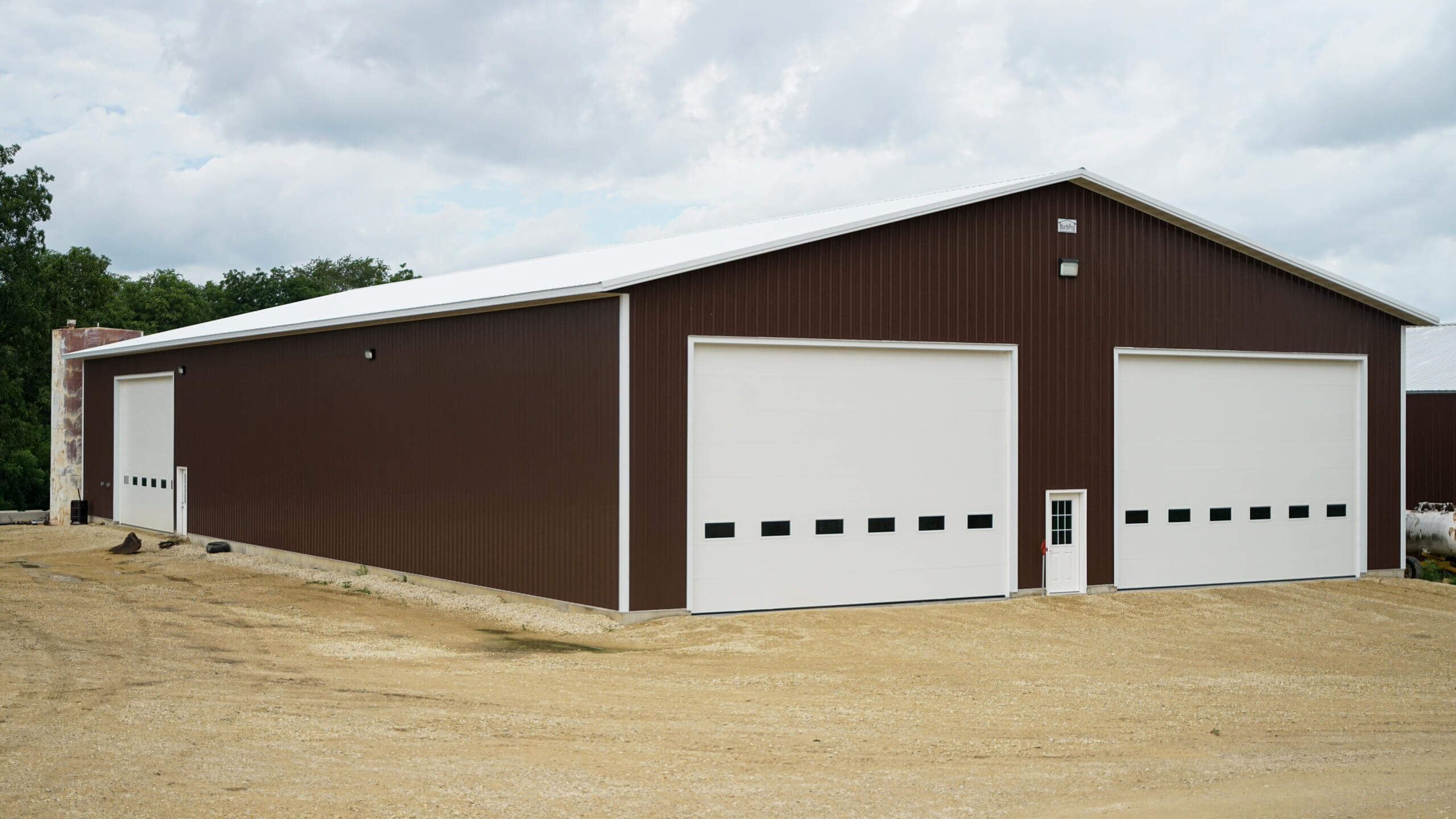 Large brown metal warehouse with two big white garage doors and a small white entry door, set on a dirt lot under a cloudy sky.