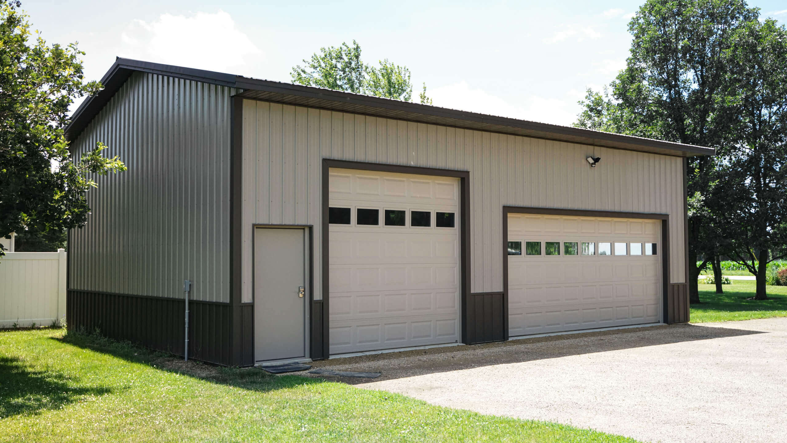 A metal garage with two large overhead doors and a single entry door, surrounded by grass and trees on a sunny day.