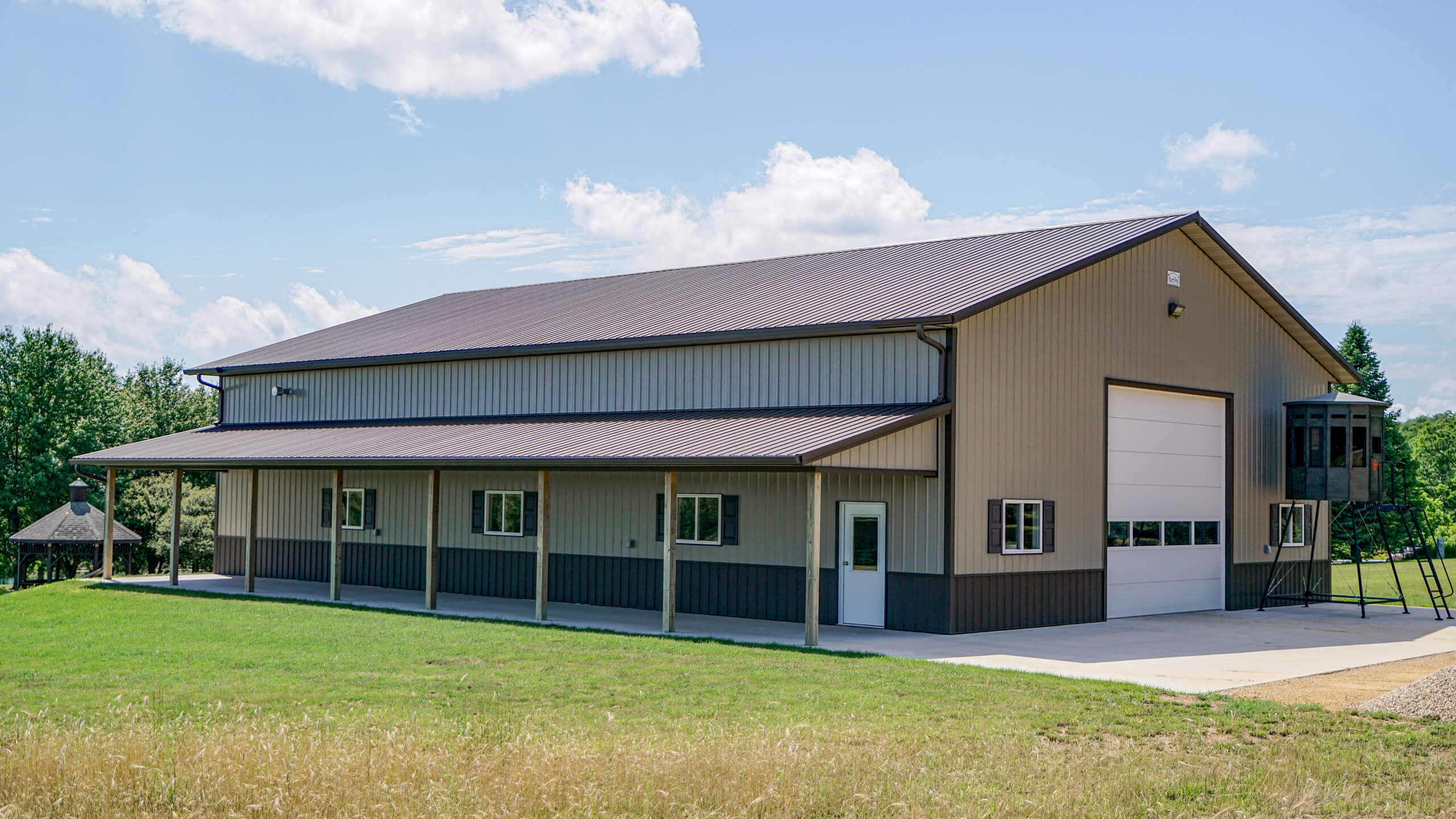 A large metal building with beige and dark brown siding, multiple windows, a garage door, and a covered walkway, set on a grassy lot under a blue sky.