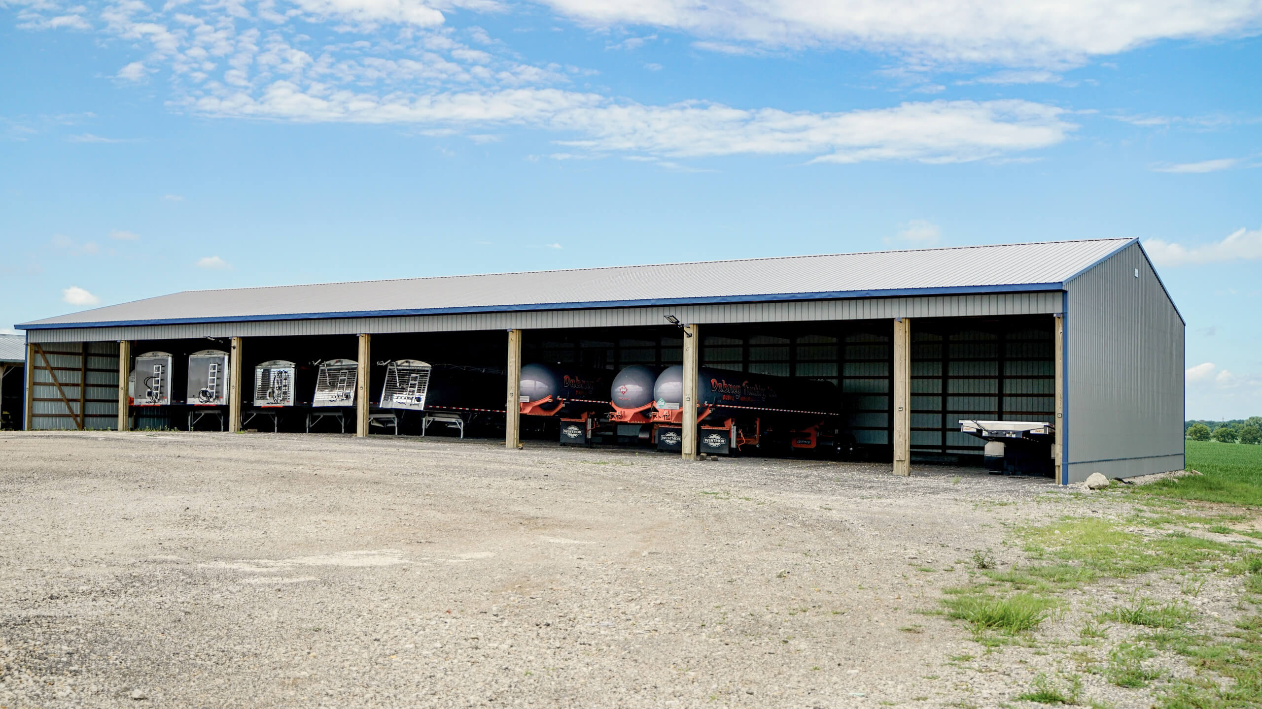Large metal shed with open front, housing several pieces of farm equipment including trailers and tanks, on a gravel lot under a partly cloudy sky.