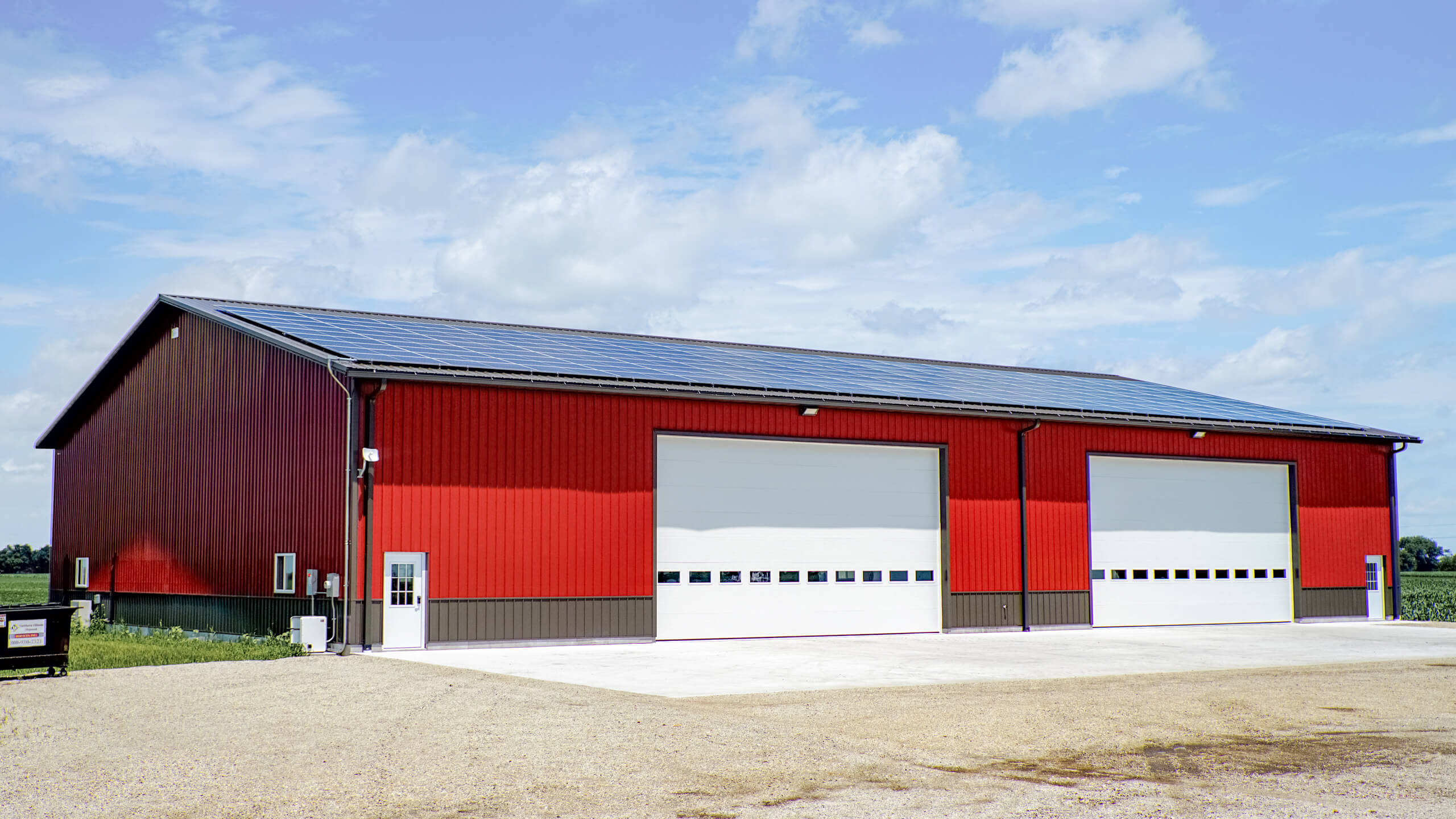 Large red metal building with three white garage doors, set on a concrete lot with gravel in the foreground and fields in the background under a partly cloudy sky.