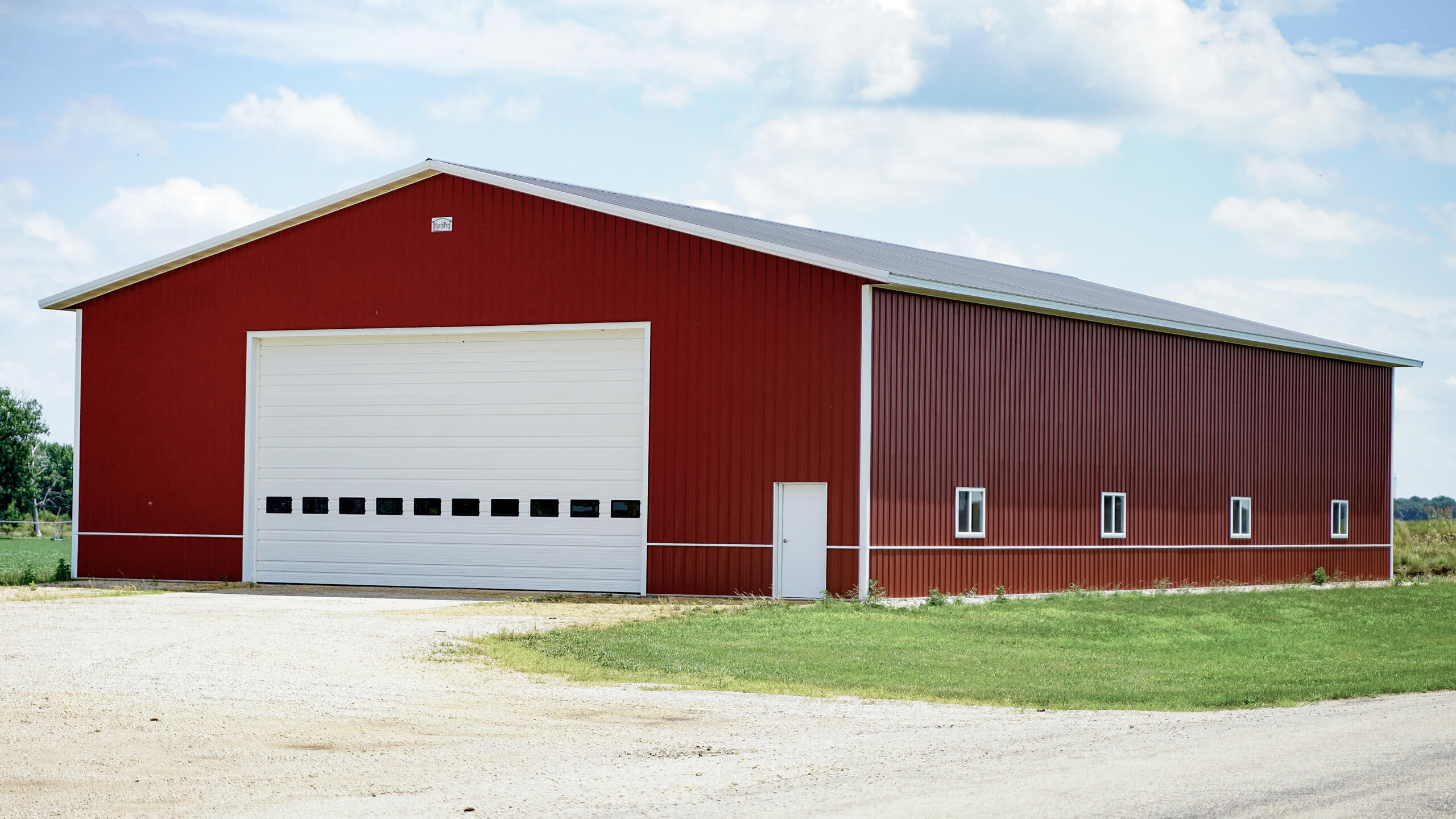 Large red metal barn with a white roof, five small windows, a white pedestrian door, and a large white garage door, situated on a grassy area under a partly cloudy sky.
