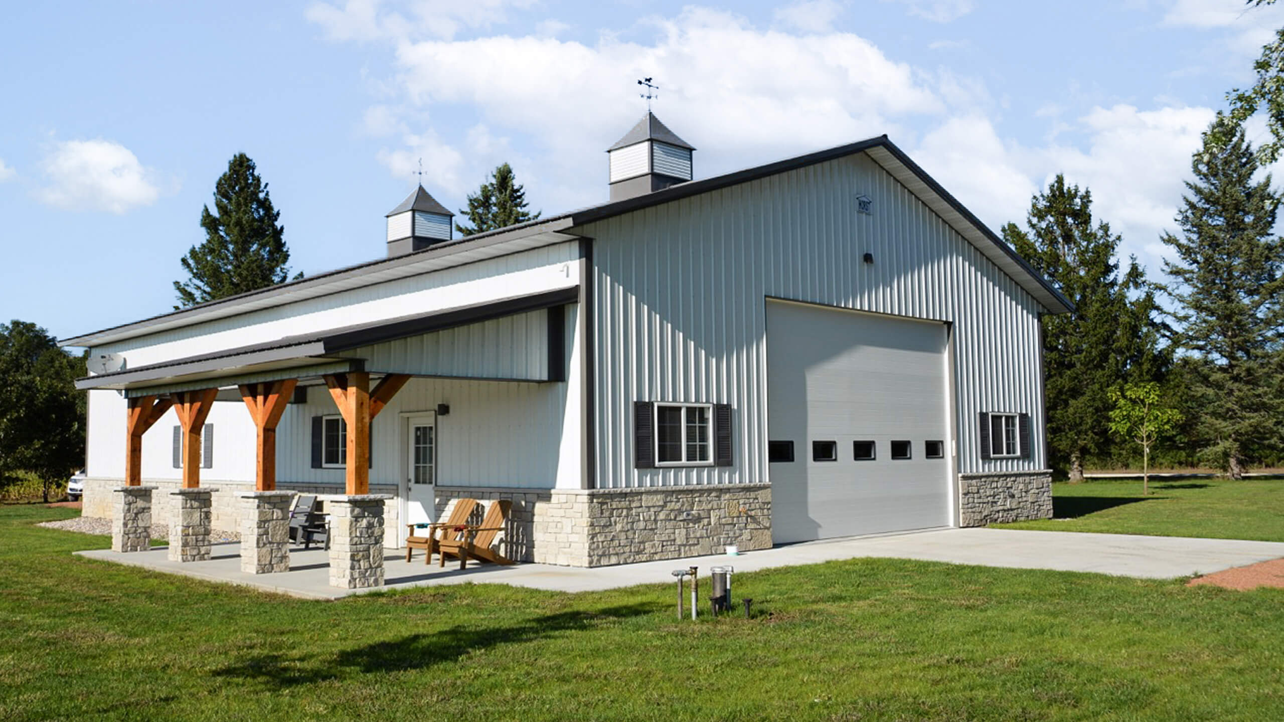 Modern metal barn with stone accents, large roll-up garage door, two cupolas, and a covered porch with chairs, set on a grassy lawn under a partly cloudy sky.