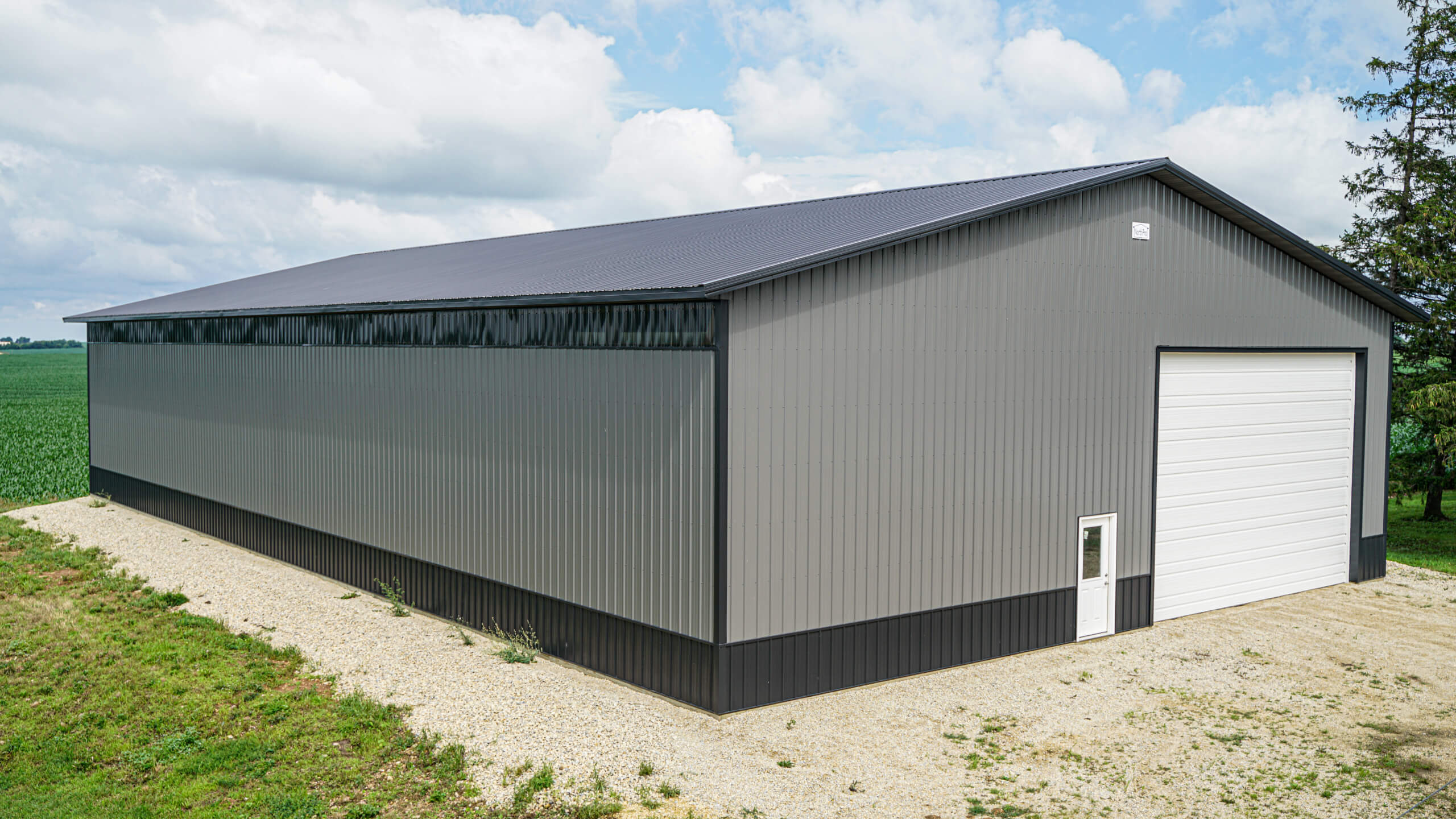 Large gray metal storage building with a black roof, white overhead door, and single entry door, situated on a gravel path beside a field and trees under a partly cloudy sky.
