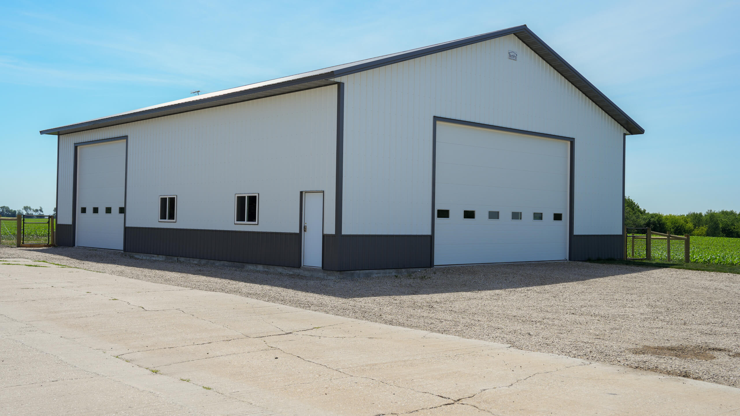 A large white metal barn with two oversized garage doors and one standard door, situated on a gravel lot next to a field under a clear blue sky.