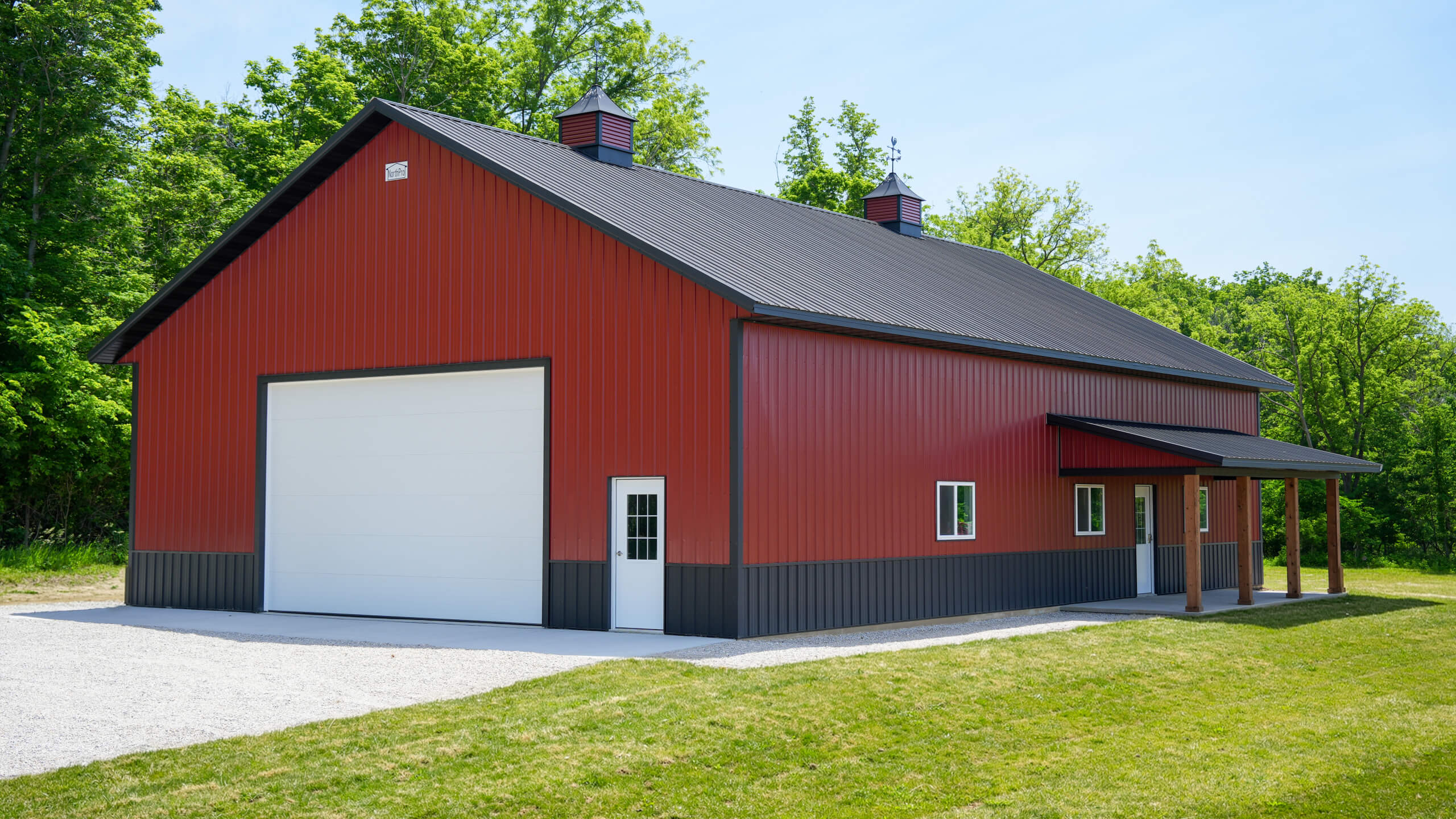Red metal barn with a large white garage door, a smaller entry door, and a covered porch, set on a grassy lot with trees in the background.