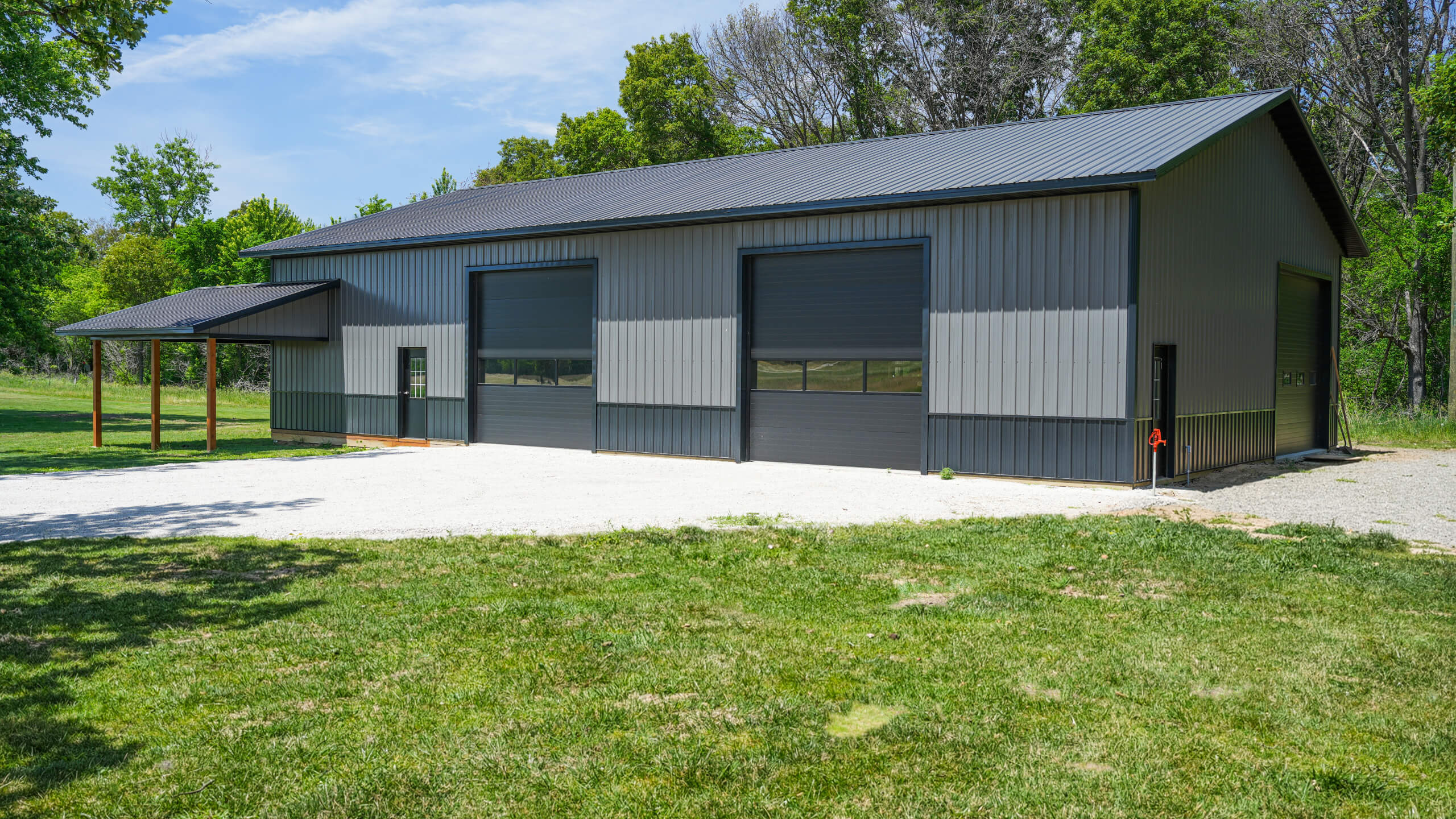 A large gray metal building with three garage doors, a side entrance, and a small covered area, set on a gravel lot surrounded by grass and trees.