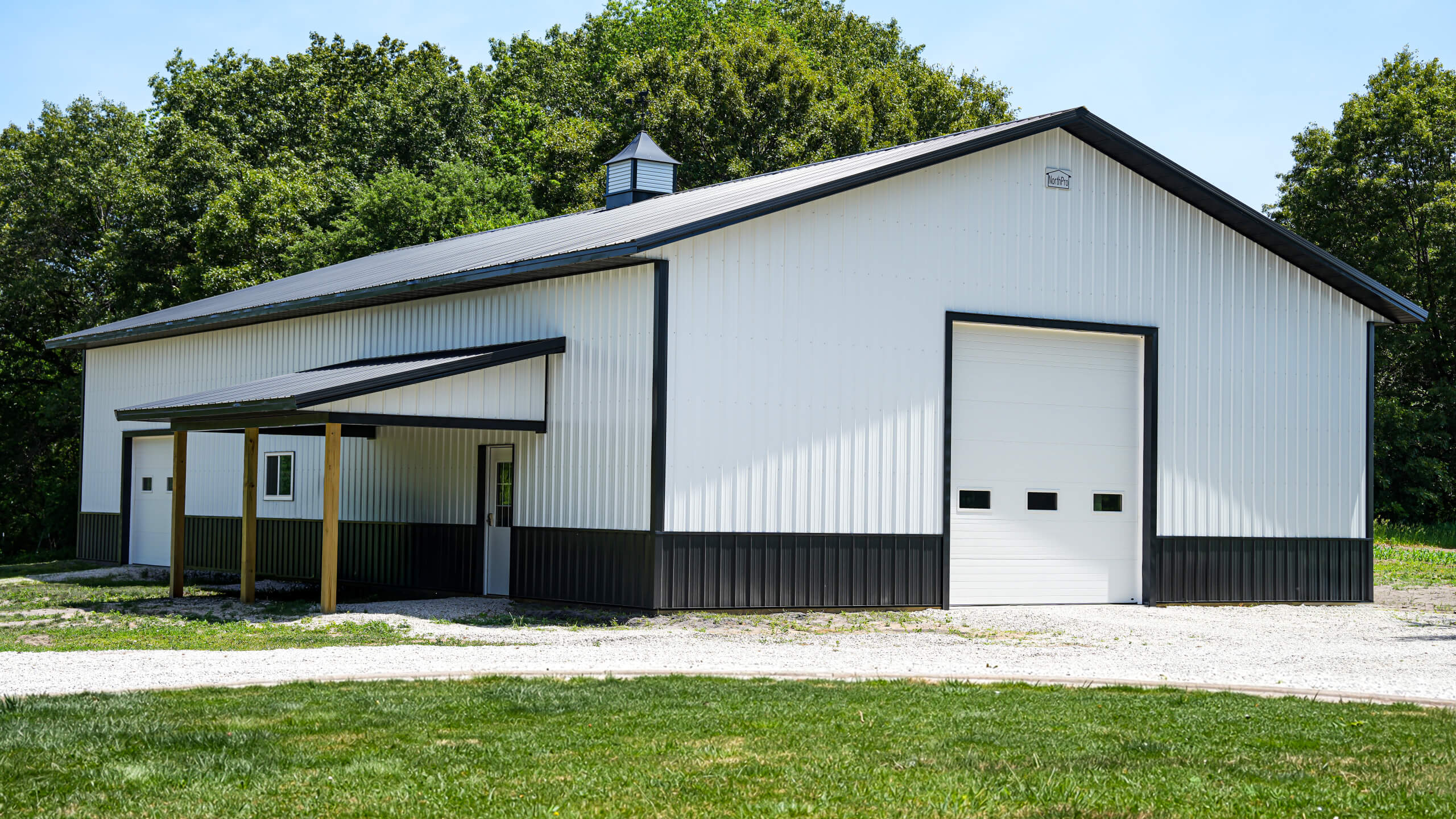 A modern white and black metal barn with a large garage door and a covered side entrance, surrounded by grass and trees.