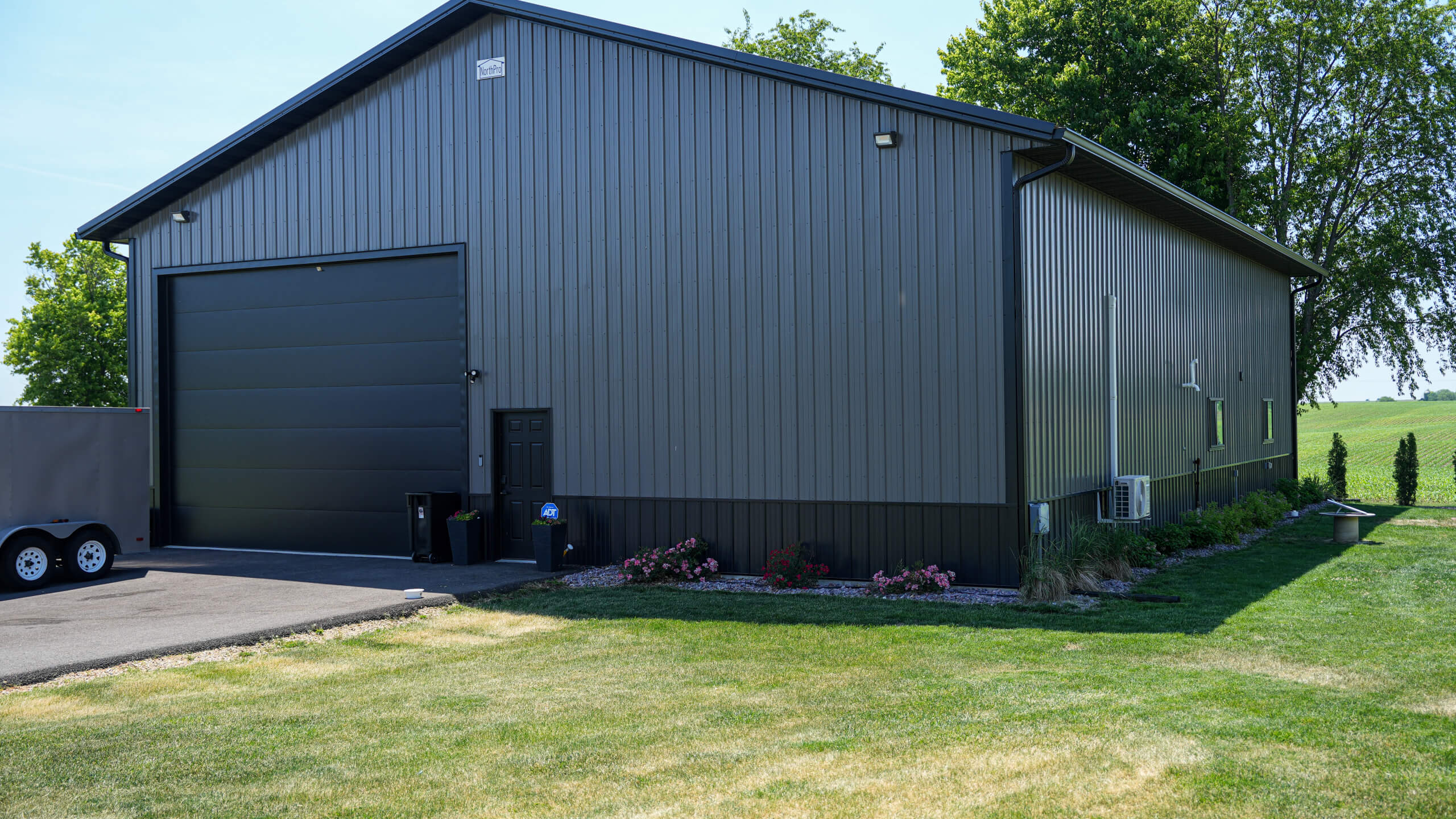 Gray metal building with a large garage door, small entry door, trailer parked nearby, landscaped flowers, and green lawn in front.