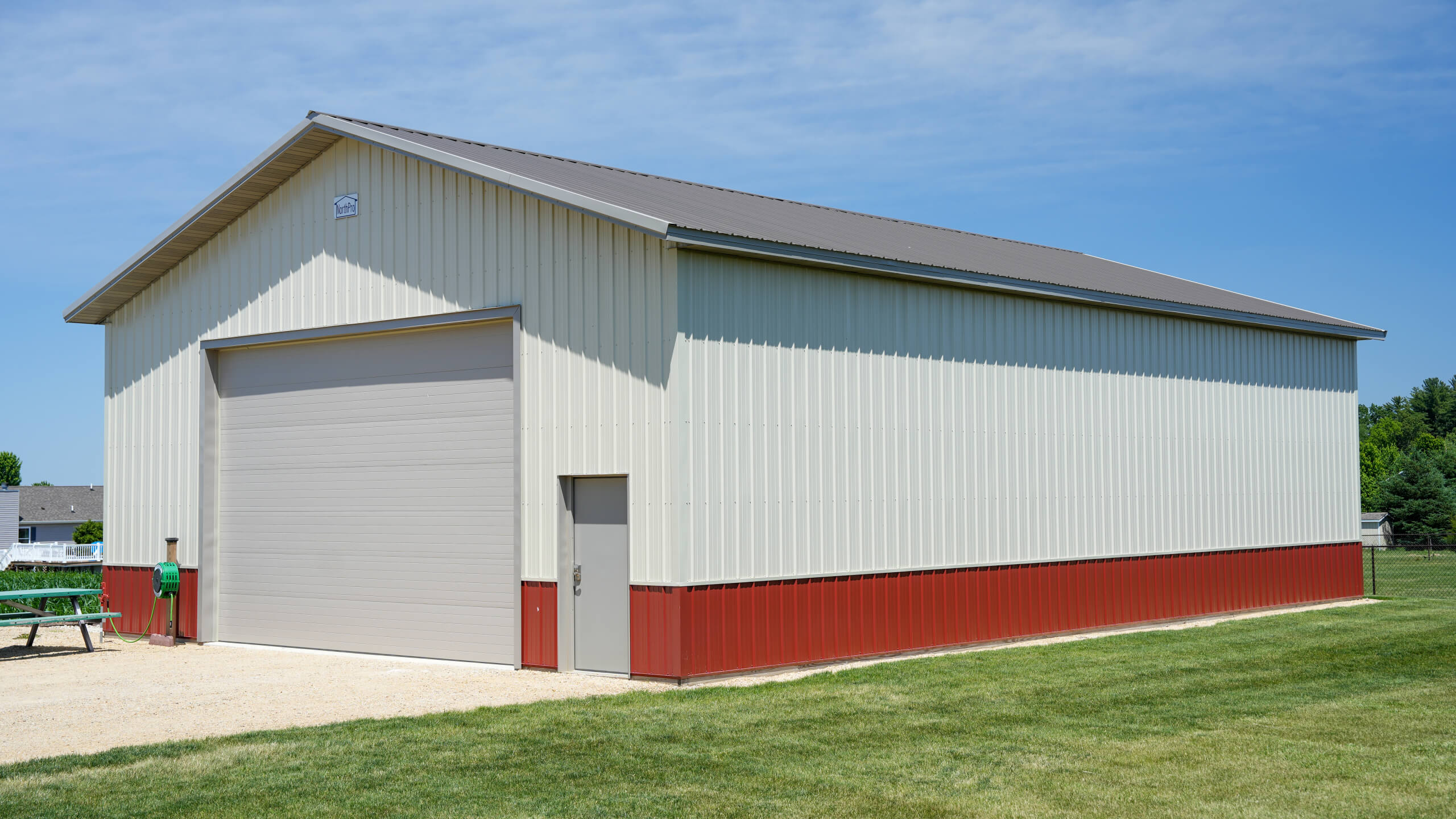 A large cream and red metal storage building with a closed garage door and small entry door, situated on a grassy area under a clear blue sky.