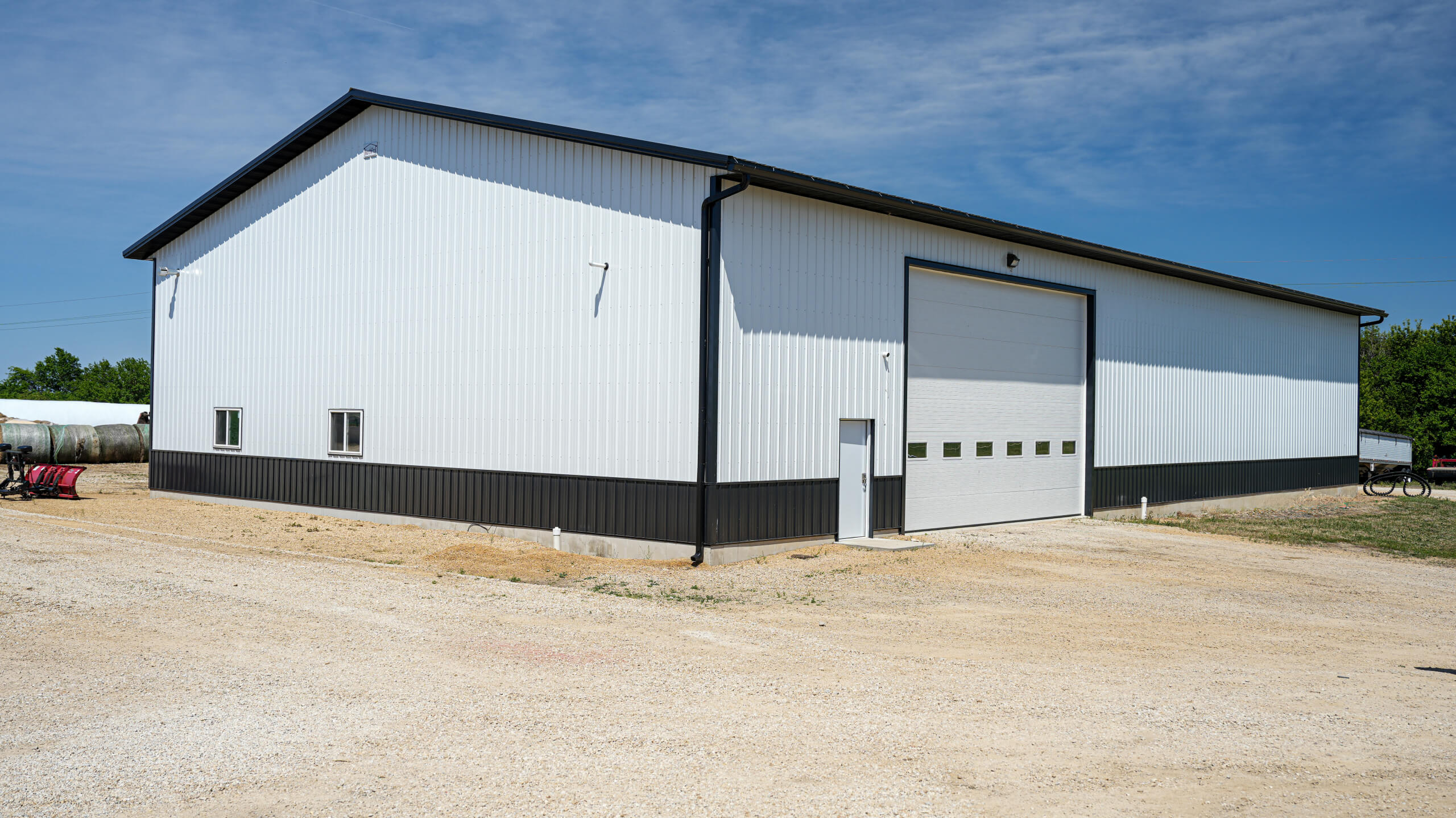 A large white metal building with a black trim and a tall garage door sits on a gravel lot under a clear blue sky.