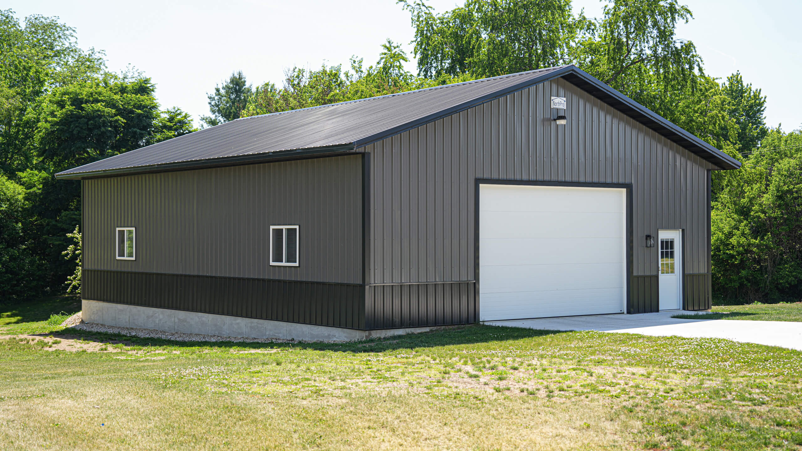 A gray metal garage building with a large white overhead door, two small windows, and a concrete driveway, surrounded by grass and trees.