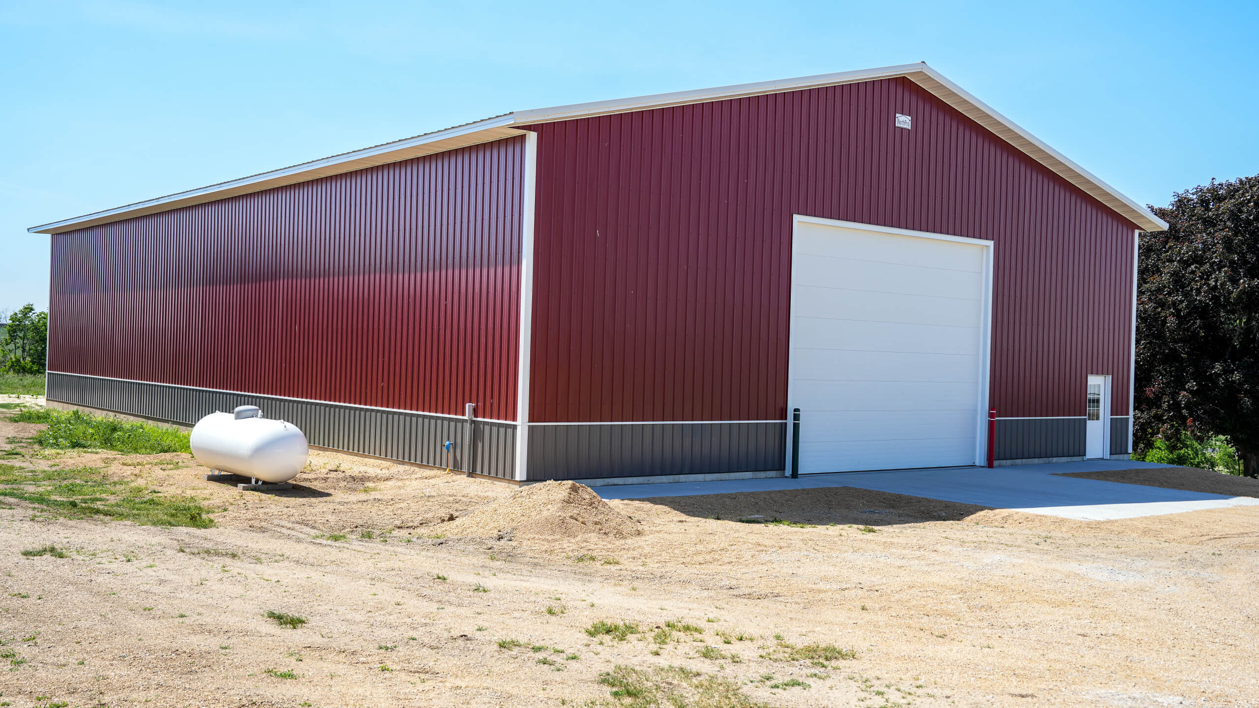 A large red metal storage building with a white garage door stands on a gravel lot. A white propane tank sits nearby. Trees are visible in the background under a clear sky.
