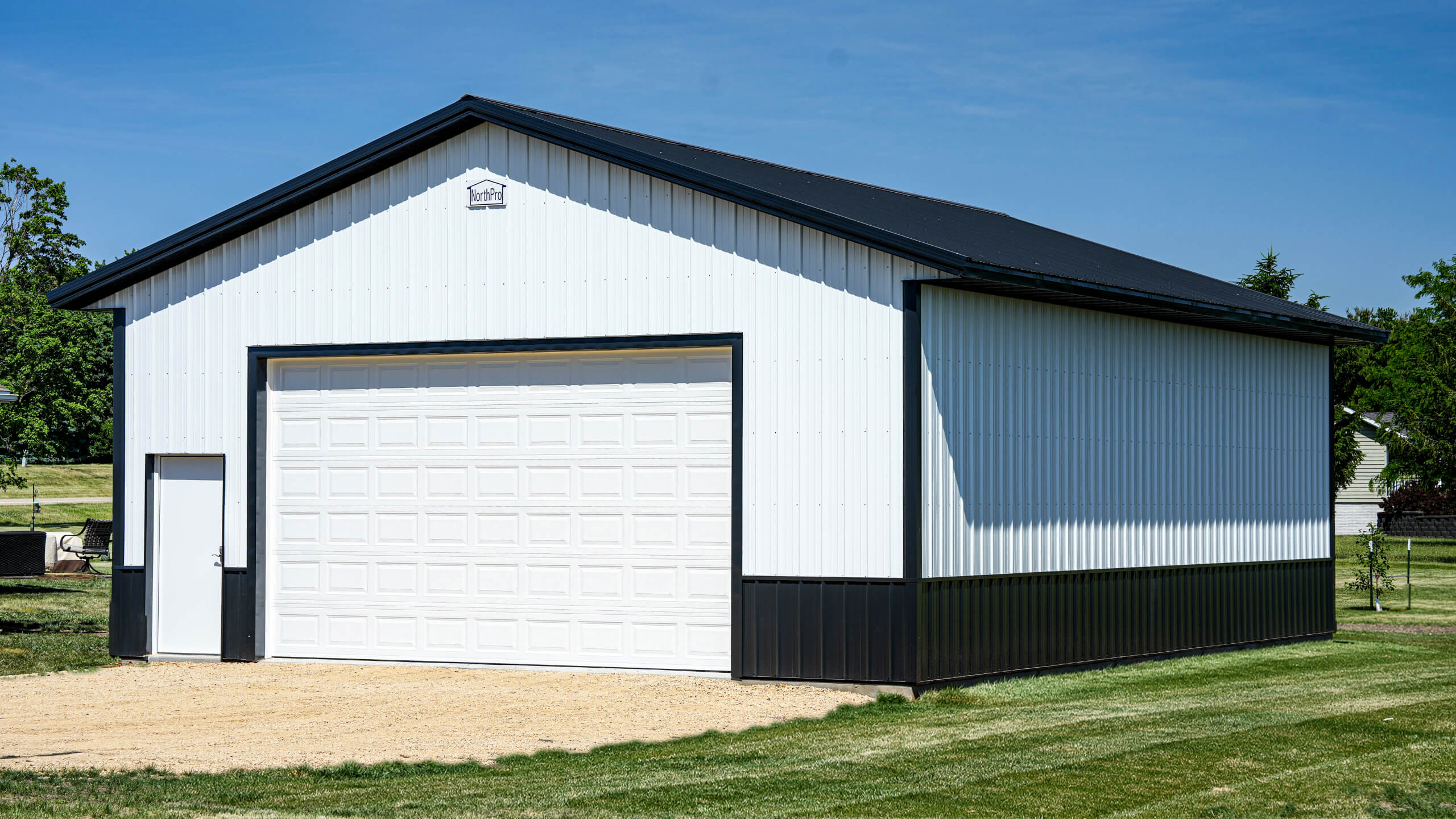 A white metal garage with a black roof and trim stands on a grassy lot under a clear blue sky.