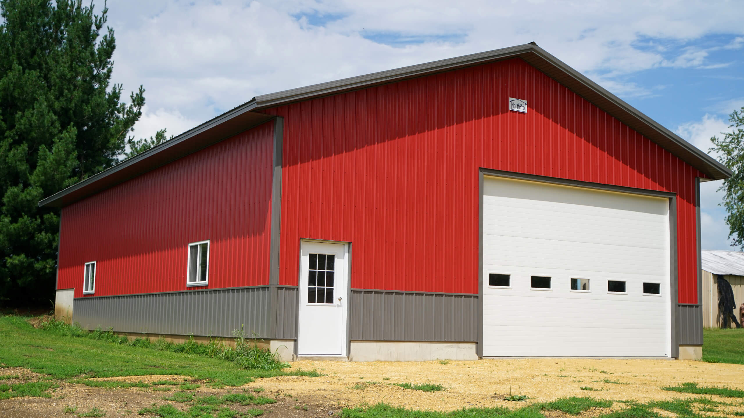 A large red and gray metal building with a white door, a row of small windows, and a wide garage door, set on a grassy area under a partly cloudy sky.