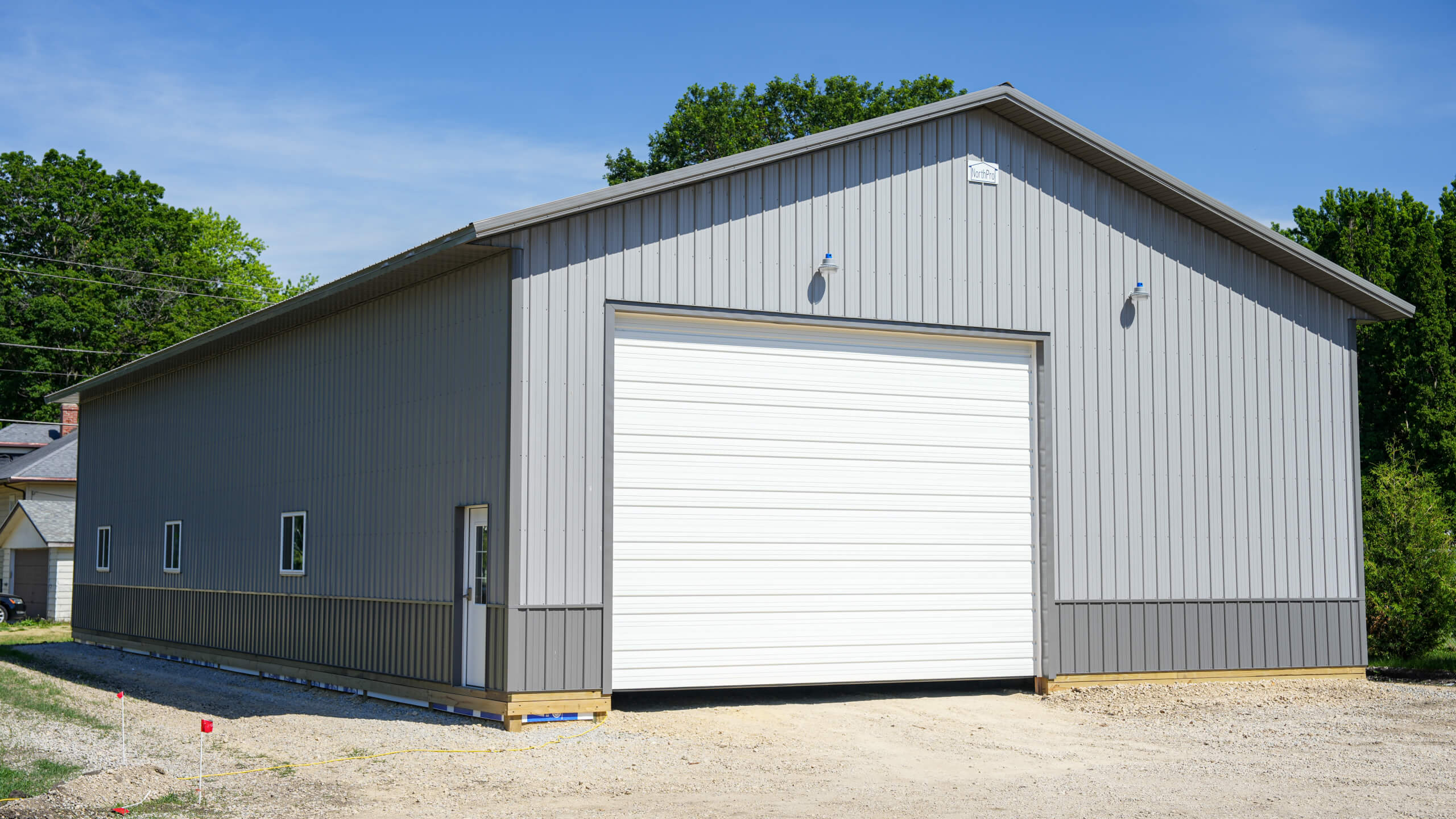 Gray metal building with a large white roll-up garage door, several small windows, and a gravel driveway in front, surrounded by green trees.