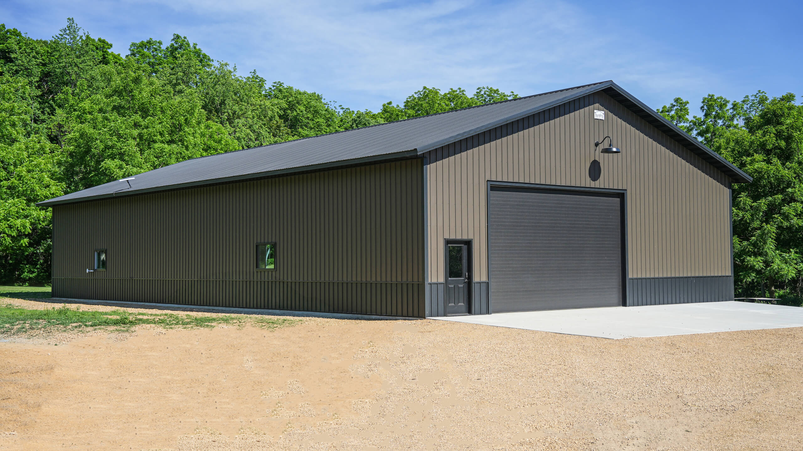 Large metal building with a black roof and roll-up garage door, surrounded by trees and a gravel driveway under a clear blue sky.