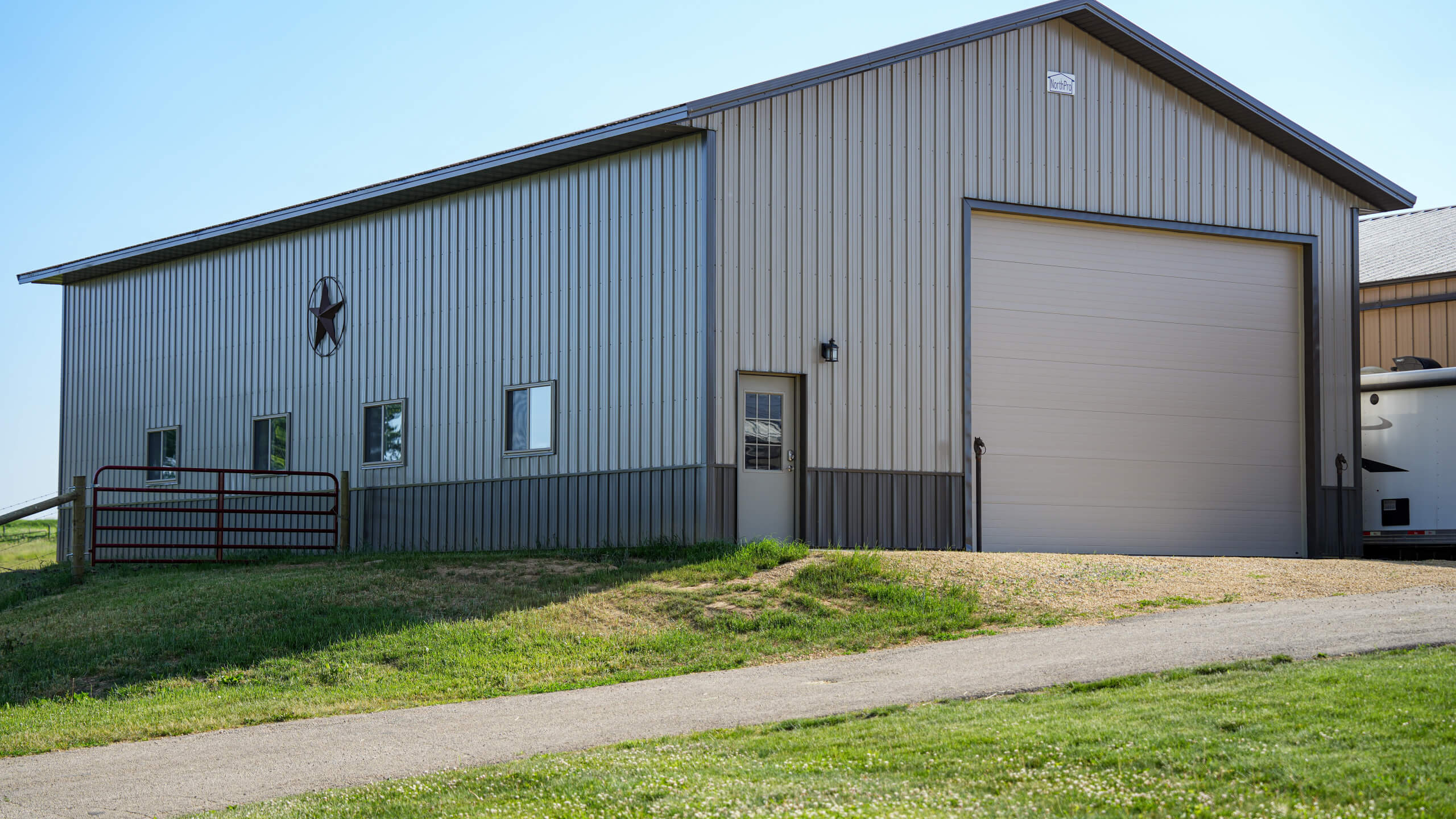 A beige metal barn with a large garage door, small windows, a star decoration on the wall, and a metal gate, set on a grassy slope next to a paved driveway.