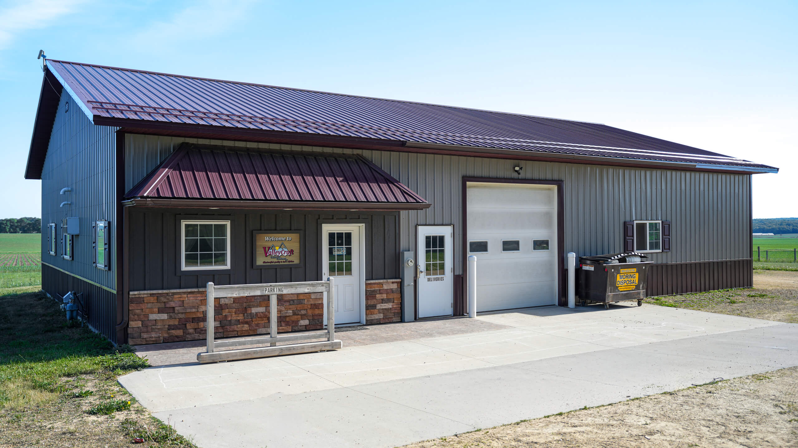 A metal building with a maroon roof, two white doors, a large garage door, and a dumpster outside, set next to a field on a sunny day.