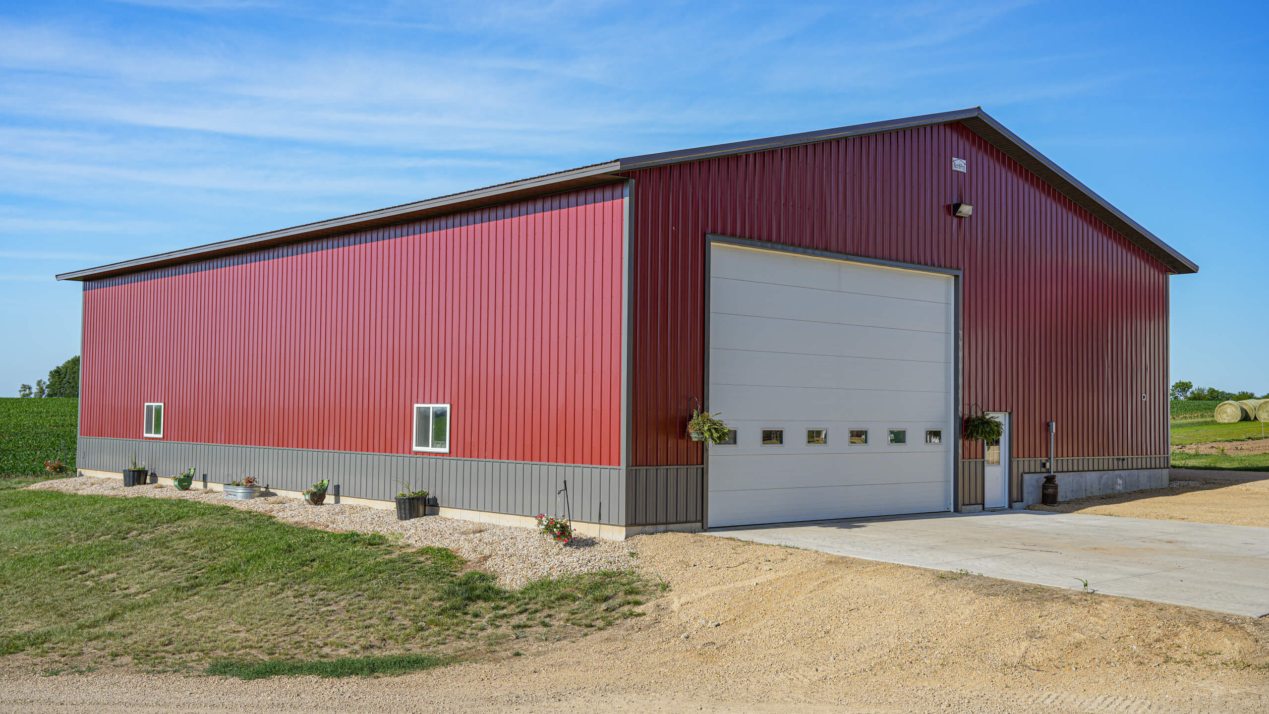 Large red metal barn with a white garage door, small windows, and potted plants in front, set on a gravel driveway beside a grassy field under a blue sky.