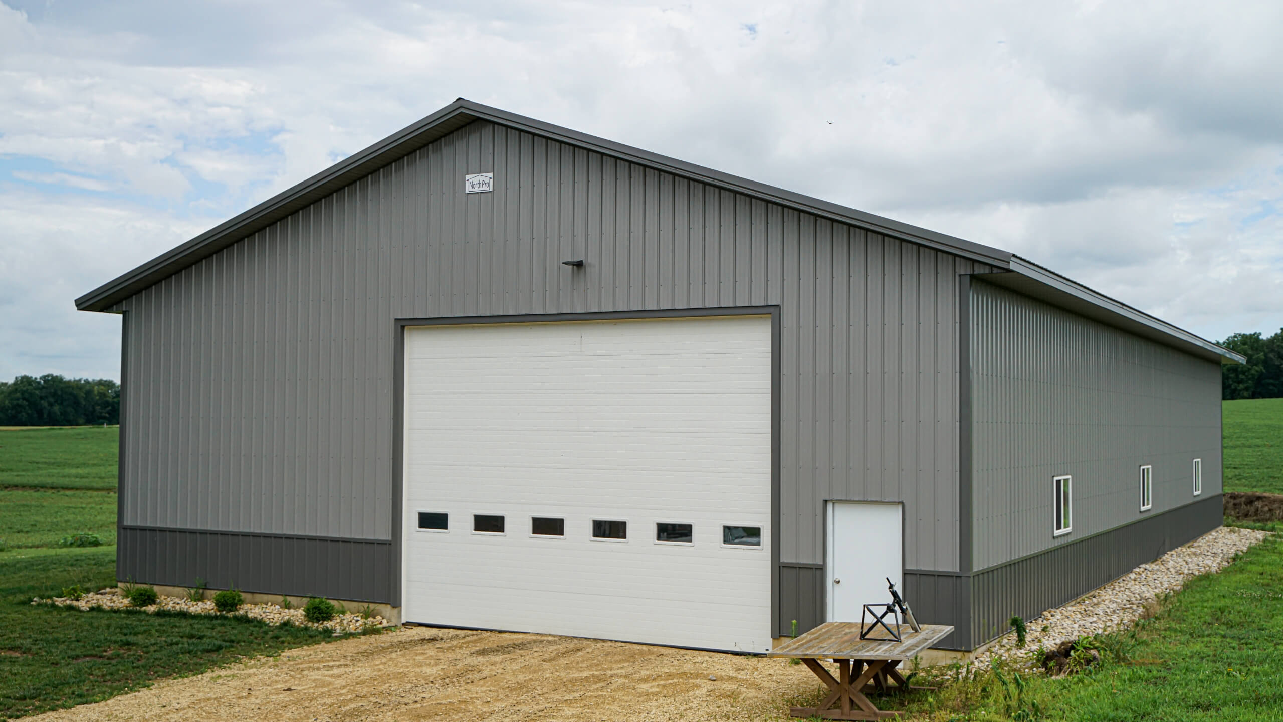 Gray metal barn with a large white garage door, small entry door, and rectangular windows, situated on a gravel path with open fields in the background.