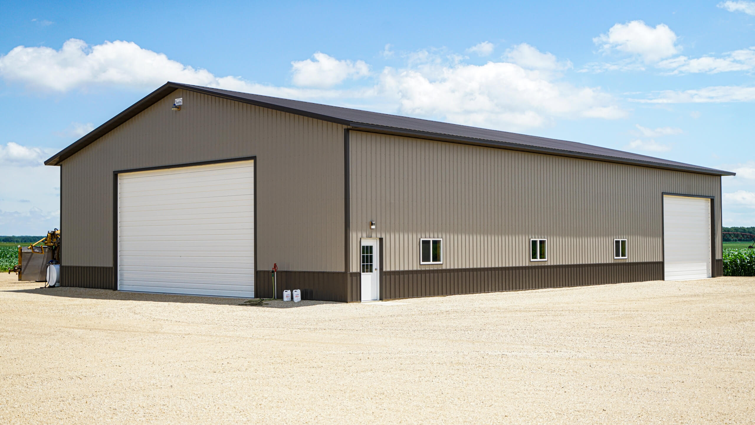Large beige metal warehouse building with two white garage doors and small windows, situated on a gravel lot under a partly cloudy sky.