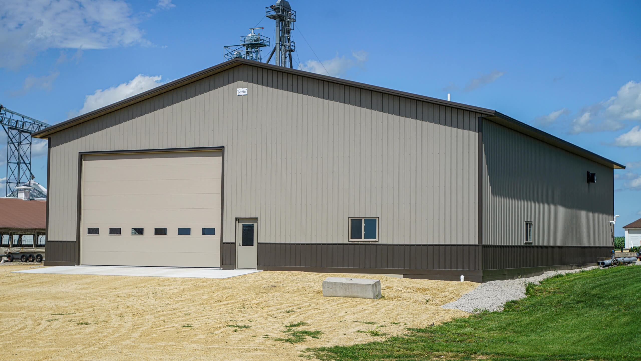 Large metal warehouse building with a beige exterior, one large garage door, a small door, and a single window, situated on a gravel lot under a clear blue sky.
