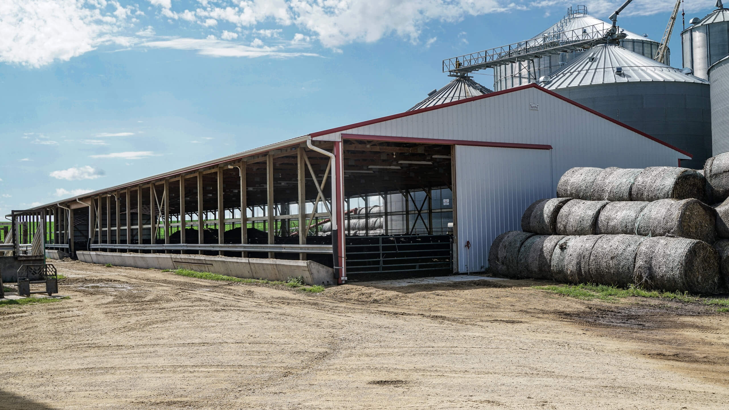 Open-sided barn with cattle inside, large stacked hay bales outside, and grain silos in the background on a farm, under a partly cloudy sky.