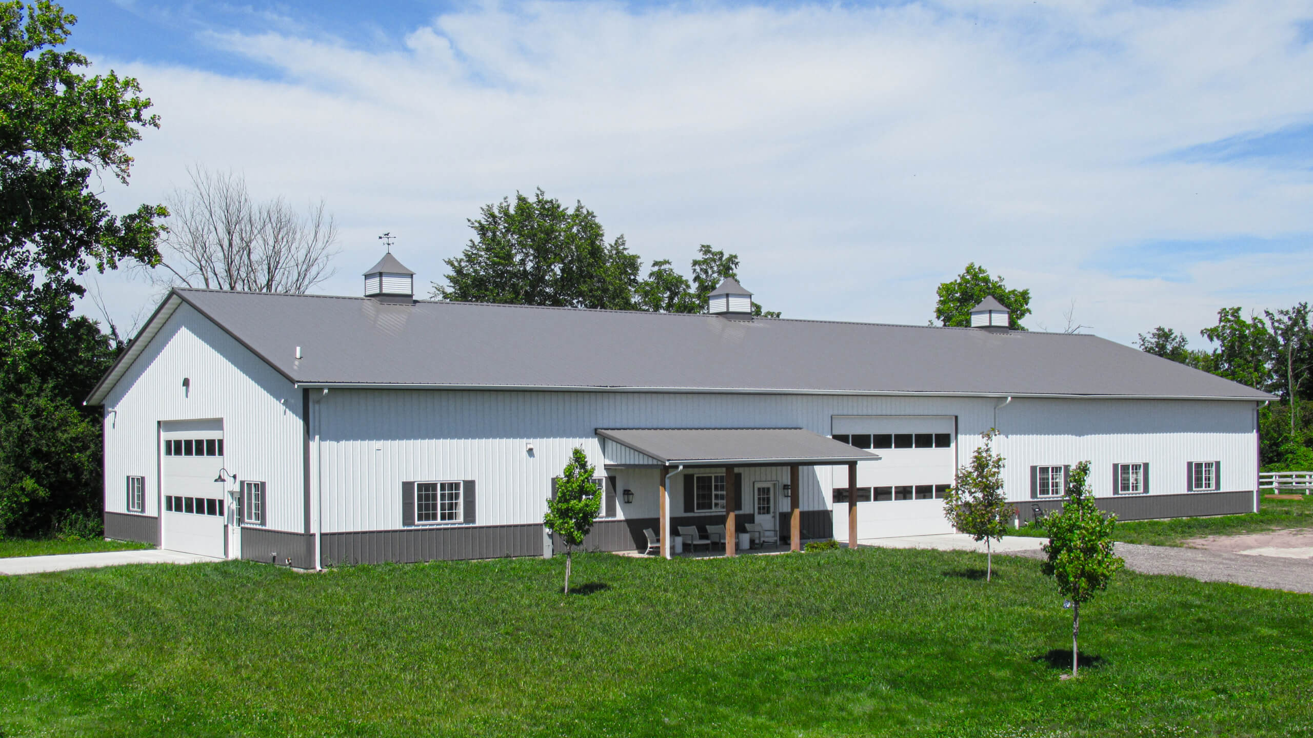 A large, white and gray metal barn with multiple garage doors and cupolas stands in a grassy area, surrounded by trees under a partly cloudy sky.