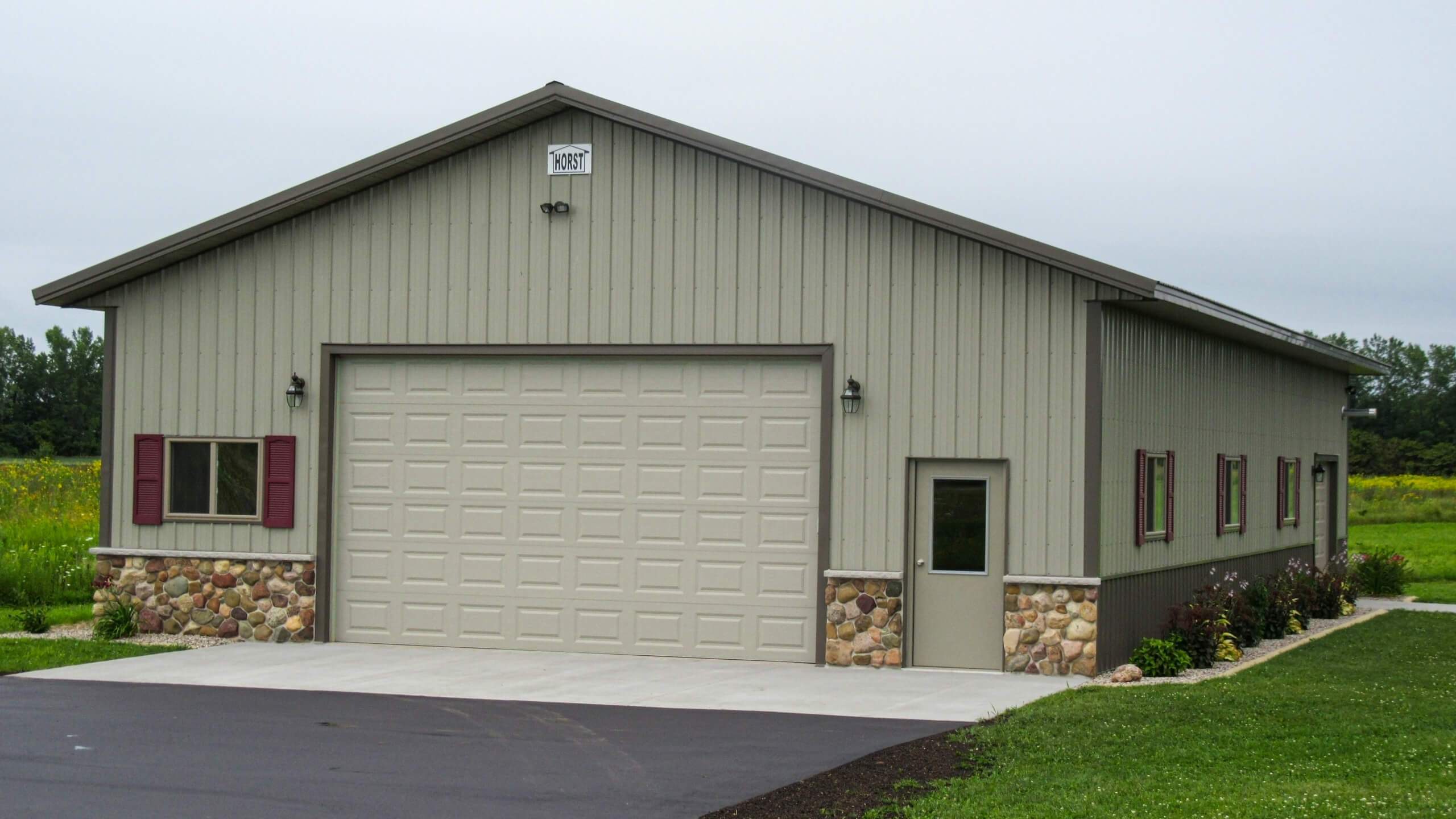 A large beige metal garage with a stone base, a wide roll-up door, a side entry door, and three windows, set on a concrete driveway beside grass.