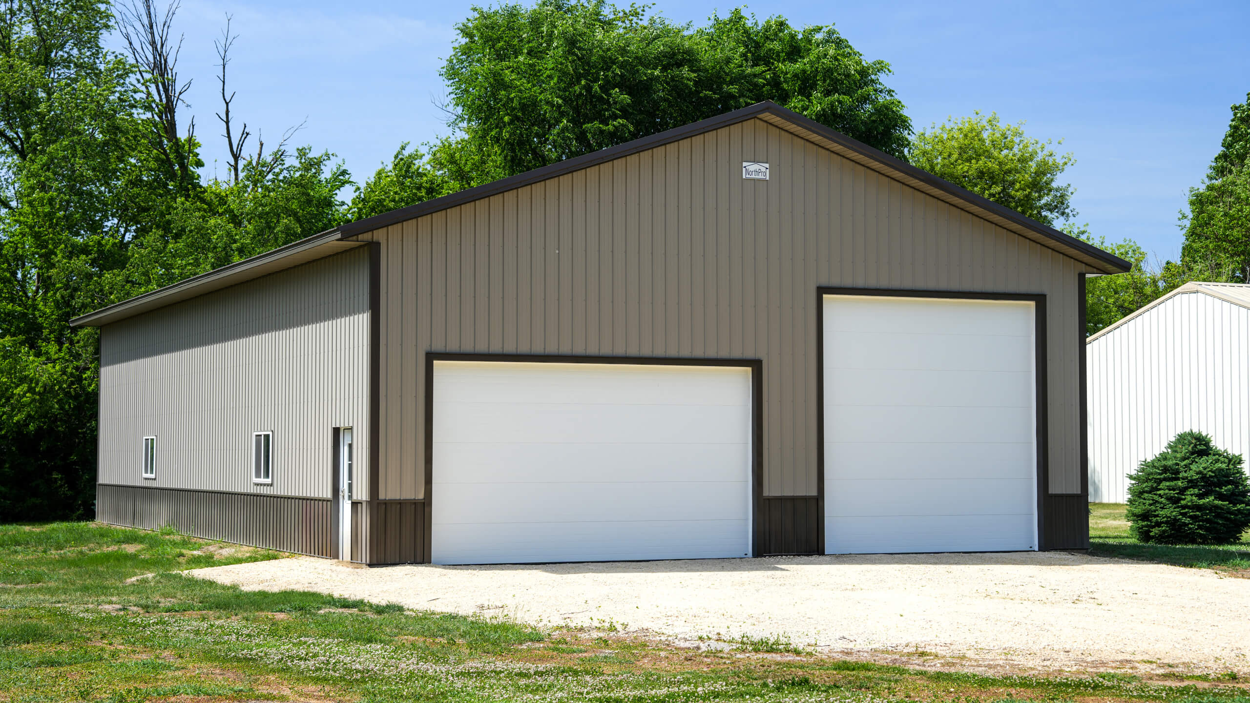 Metal storage building with two large white garage doors, set on a gravel lot, surrounded by green trees and grass on a sunny day.